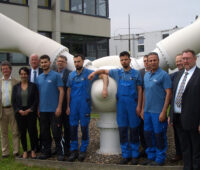 Eine Gruppe von Menschen vor dem Kopf eines im Boden verankerken relativ keinen Rotorkopfes einer Windkraftanlage mit Flügeln