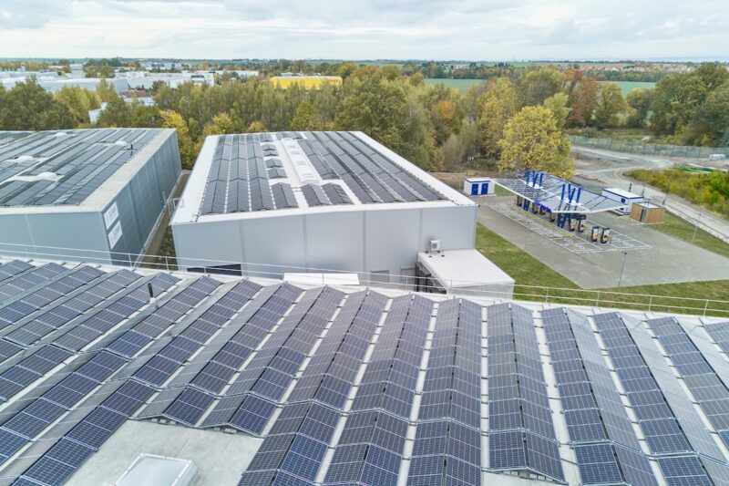 Im Vordergrund industrielle Hallendächer mit Photovoltaikanlage - hinten rechts befindet sich die Schnelladestation für E-Autos.