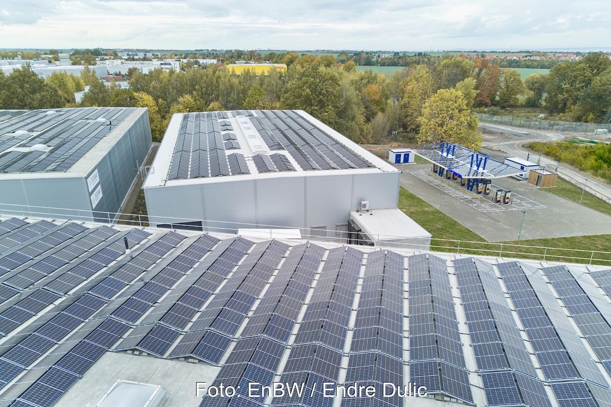 Im Vordergrund industrielle Hallendächer mit Photovoltaikanlage - hinten rechts befindet sich die Schnelladestation für E-Autos.