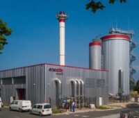 Gebäude einer Energiezentrale unter blauem Himmel und historischem Wasserspeicher im Hintergrund.