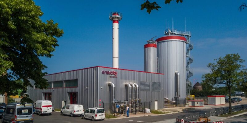 Gebäude einer Energiezentrale unter blauem Himmel und historischem Wasserspeicher im Hintergrund.