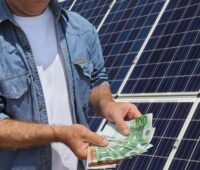 Zu sehen ist ein Mensch mit Geldscheinen vor einer Photovoltaik-Anlage - Symbol für PV-Miete im Vergleich zum Kauf.