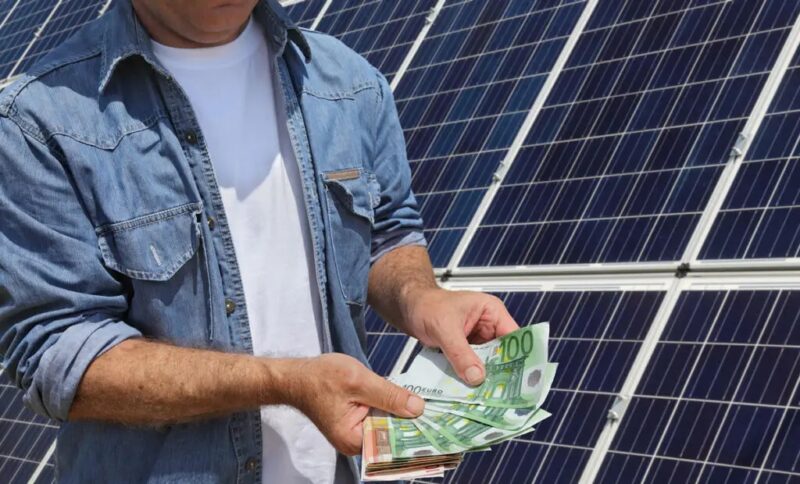 Zu sehen ist ein Mensch mit Geldscheinen vor einer Photovoltaik-Anlage - Symbol für PV-Miete im Vergleich zum Kauf.