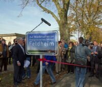 Viele Menschen hinter einem Eingangschild mit Foto des Dorfes und der Aufschrift: Auf Wiedersehen im Energieautarken Ortsteil Feldheim.
