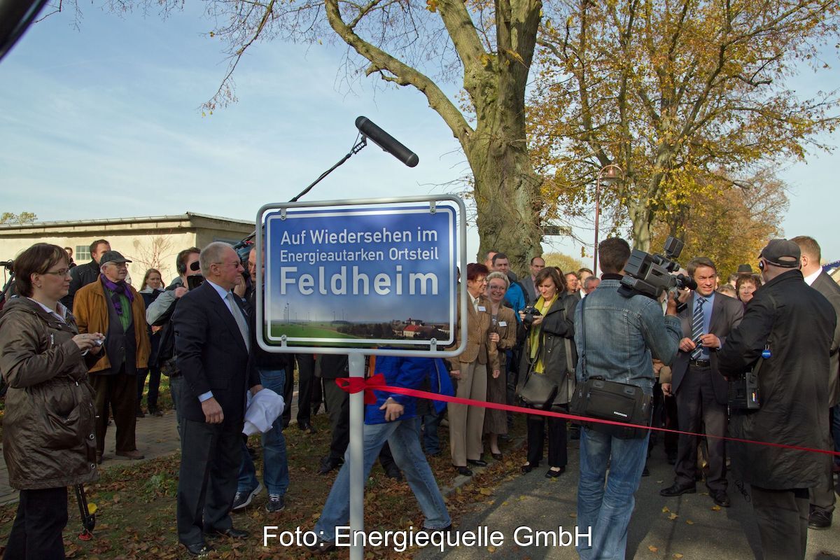 Viele Menschen hinter einem Eingangschild mit Foto des Dorfes und der Aufschrift: Auf Wiedersehen im Energieautarken Ortsteil Feldheim.
