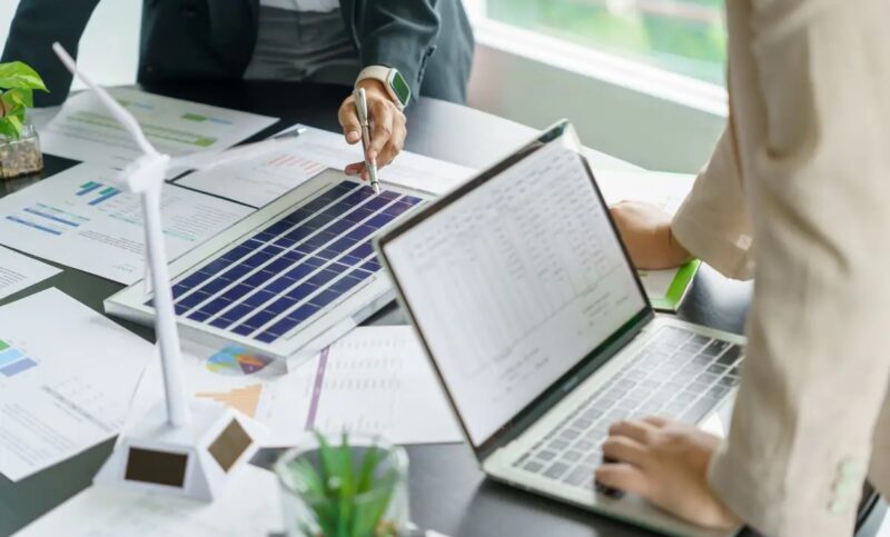 Zwei Personen an einem Tisch mit Laptop, Windradmodell und PV-Modell, der Wirtschaftlichkeitsrechner für Home Energy Management Systeme von Energielenker berechnet Szenarien.