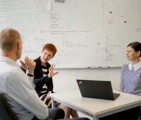 Ein Mann und eine Frau gestikulierend im Gespräch, eine junge Frau sitzt hinter einem Computer - Forschungsgruppe für Energiewende-Kommunikation.