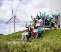 Menschengruppe zeigt am Fuß einer WIndenergieanlage Plakate und ähnliches, um für Bürgerenergie und Energy Sharing zu demonstrieren.