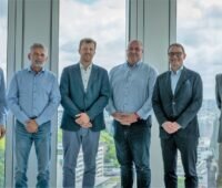 Sechs Männer im leichten Business-Outfit vor einem Fenster
