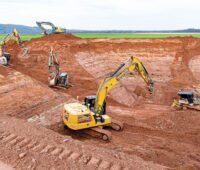 Baugrube der Erdbecken-Wärmespeichers - Saisonalspiechers - in Bracht. Vier Bagger und ein Bulldozer arbeiten in rotem Gestein.