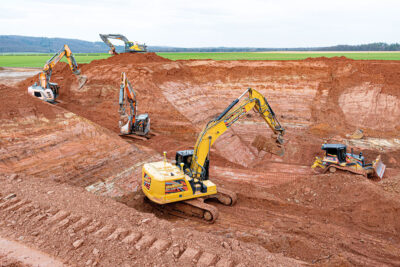 Baugrube der Erdbecken-Wärmespeichers - Saisonalspiechers - in Bracht. Vier Bagger und ein Bulldozer arbeiten in rotem Gestein.