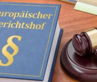 Im Bild ein blaues Buch mit Paragraphenzeichen und der Aufschrift Europäischer Gerichtshof, daneben ein hölzerner Richterhammer, beides liegt auf einer Tischplatte.