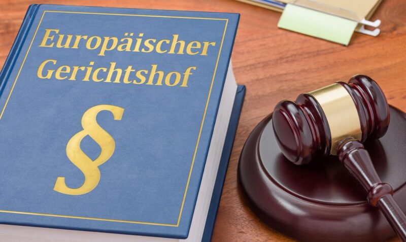 Im Bild ein blaues Buch mit Paragraphenzeichen und der Aufschrift Europäischer Gerichtshof, daneben ein hölzerner Richterhammer, beides liegt auf einer Tischplatte.