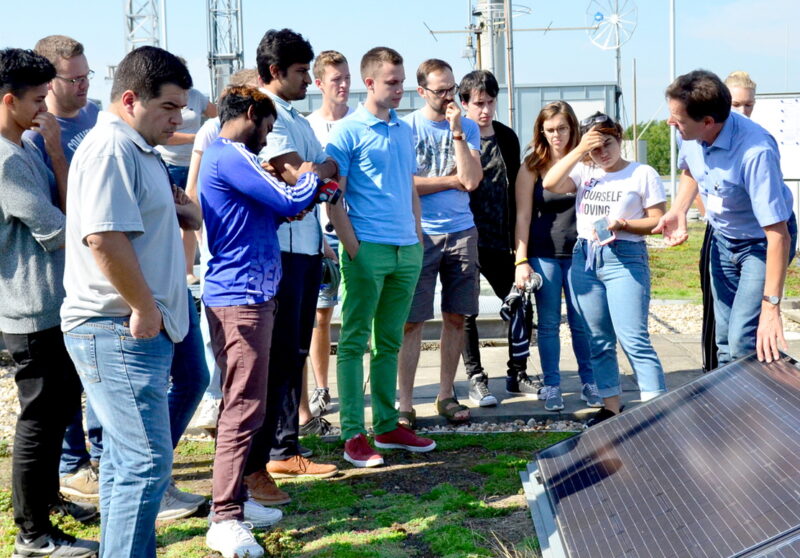 Studenten hören im Freien einem Lehrer zu, der an einem Photovoltaikmodul steht.