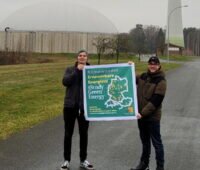 Zwei junge Männer halten ein Plakat, das über Studienmöglichkeiten informiert. Im HIUntergrund ein Biogasfermenter.