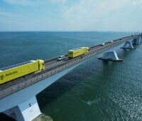 Auf dem Foto ist eine weitläufige Brücke über ein Meer zu sehen, die von links unten schräg nach rechts oben in den Horizont läuft. Auf der Brücke fahren neben PKWs zwei gelbe Trucks mit PV auf dem Dach.