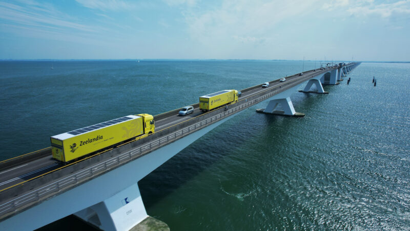Auf dem Foto ist eine weitläufige Brücke über ein Meer zu sehen, die von links unten schräg nach rechts oben in den Horizont läuft. Auf der Brücke fahren neben PKWs zwei gelbe Trucks mit PV auf dem Dach.