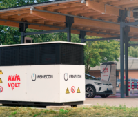 Weiß-schwarzer FENECON-Batteriespeicher im Vordergrund eines überdachten Ladeparks. Dahinter steht ein Auto an zwei AVIA-Volt-Schnellladesäulen.