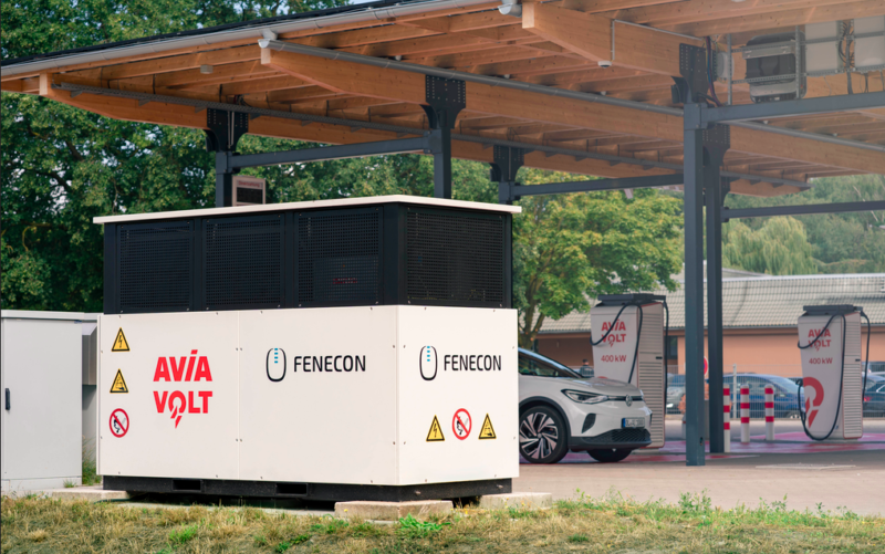Weiß-schwarzer FENECON-Batteriespeicher im Vordergrund eines überdachten Ladeparks. Dahinter steht ein Auto an zwei AVIA-Volt-Schnellladesäulen.