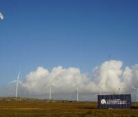 Ein Drachen fliegt vor Windenergieanlagen, Kitepower testet sein Flugwindkraft-System in Irland.