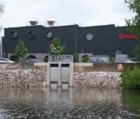 Das Bild zeigt von der Flussseite aus gesehen ein schwarzes Gebäude mit den drei roten Buchstaben SWG an der Seite. An der zwischen Gebäude und Fluss liegenden Straße parken Autos.