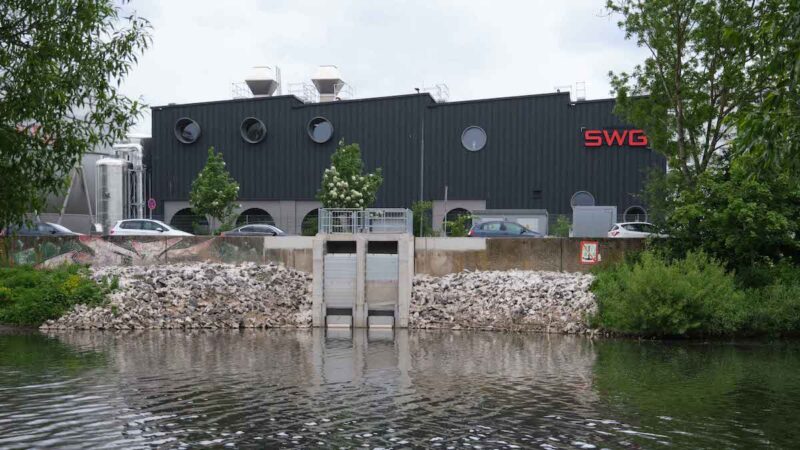 Das Bild zeigt von der Flussseite aus gesehen ein schwarzes Gebäude mit den drei roten Buchstaben SWG an der Seite. An der zwischen Gebäude und Fluss liegenden Straße parken Autos.
