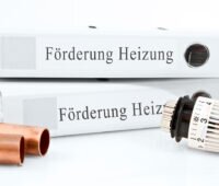 Symbolfoto für Förderung der Heizungserneuerung. Auf weißem Untergrund: zwei Ordner mit Aufschrift "Förderung Heizung" Kupferrohre und ein Thermostatventilkopf.