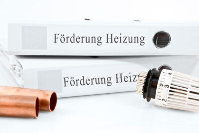 Symbolfoto für Förderung der Heizungserneuerung. Auf weißem Untergrund: zwei Ordner mit Aufschrift "Förderung Heizung" Kupferrohre und ein Thermostatventilkopf.