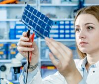 Eine Frau hält eine Folie mit kleinen Solarzellen in die Höhe, zwei Klemmen sind daran angebracht - Energieforschung, Photovoltaik. Dünnschicht