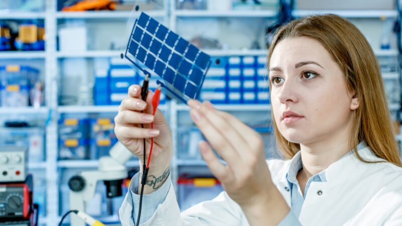 Eine Frau hält eine Folie mit kleinen Solarzellen in die Höhe, zwei Klemmen sind daran angebracht - Energieforschung, Photovoltaik. Dünnschicht