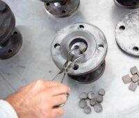 Eine Hand hält eine Zange und legt eine graue Kugel in einen Stahlzylinder - Wärmespeicher-Experiment am Fraunhofer ISE.