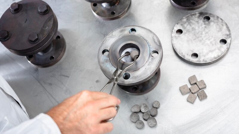 Eine Hand hält eine Zange und legt eine graue Kugel in einen Stahlzylinder - Wärmespeicher-Experiment am Fraunhofer ISE.