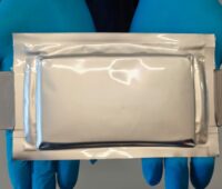 Kunststoff ummantelte Batteriezelle präsentiert von zwei Händen in Handschuhen.