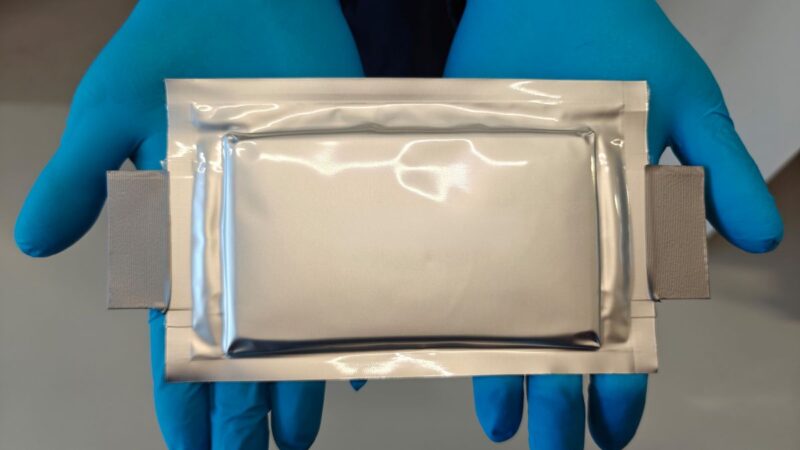 Kunststoff ummantelte Batteriezelle präsentiert von zwei Händen in Handschuhen.