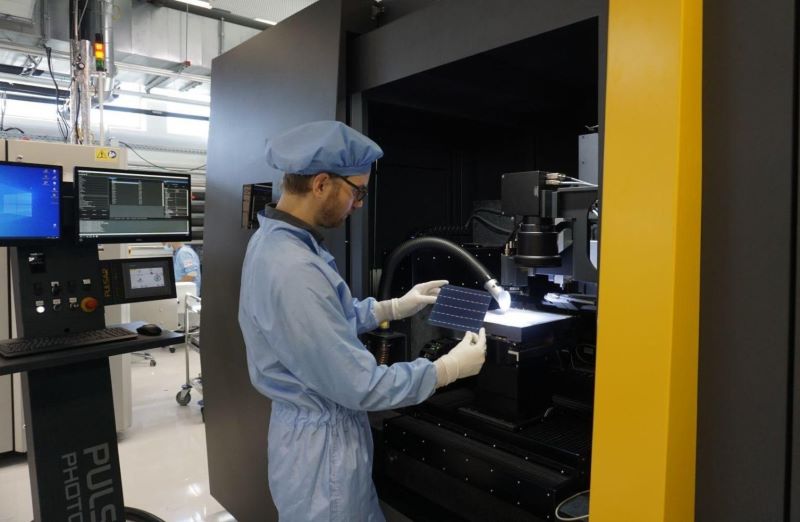 Ein Laborant hält eine Solarzelle vor einer Maschine in der Hand.