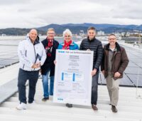 Fünf Personen auf einem Stadiondach mit großer Urkunde vor PV-Anlage.