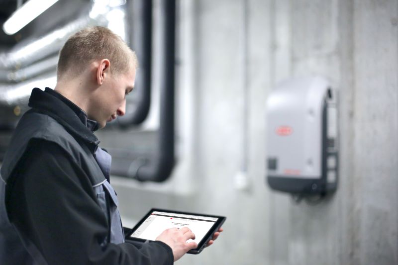 Ein Techniker blickt vor dem Hintergrund eines Wechselrichters an der Wand auf ein Tablet.