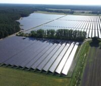 Der Solarpark Frankenförde, geteilt durch einen Hain, zwischen Feldern; links davon ein Waldgebiet. Das Sonnenlicht reflektiert von rechts auf den Modulen.