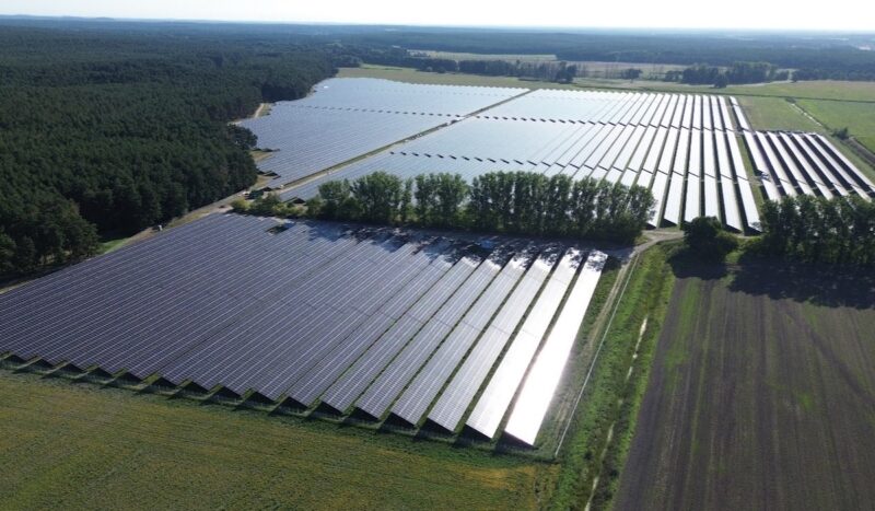 Der Solarpark Frankenförde, geteilt durch einen Hain, zwischen Feldern; links davon ein Waldgebiet. Das Sonnenlicht reflektiert von rechts auf den Modulen.