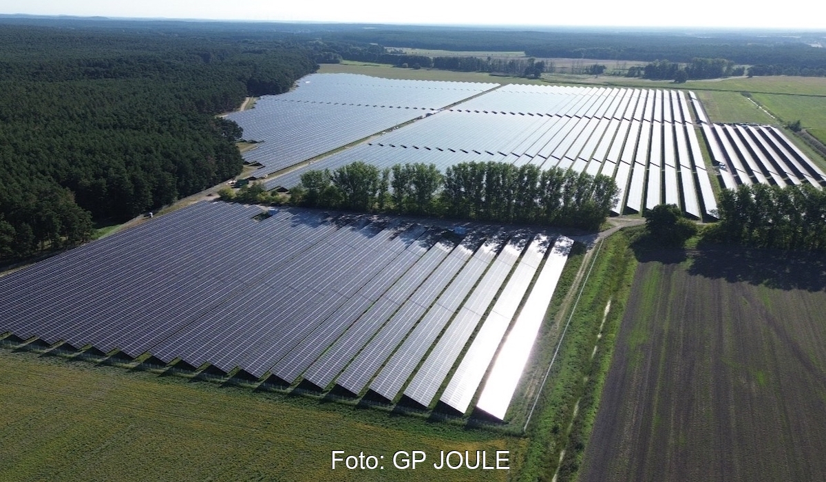 Der Solarpark Frankenförde, geteilt durch einen Hain, zwischen Feldern; links davon ein Waldgebiet. Das Sonnenlicht reflektiert von rechts auf den Modulen.