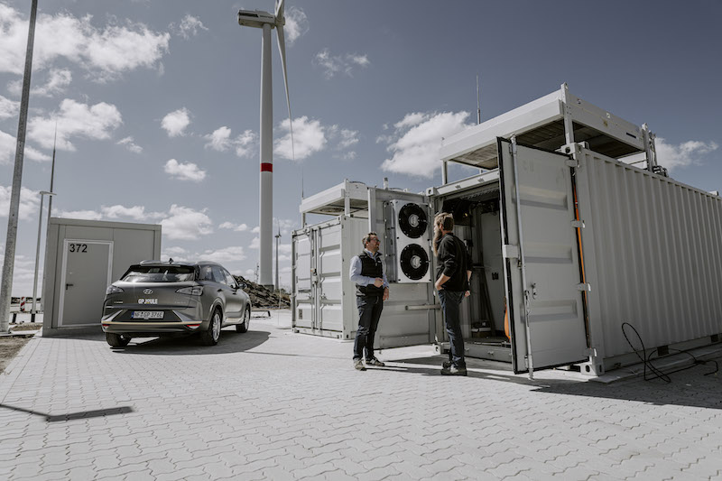 Rechts ein Container mit technischen Anlagen, im Hintergrund eine Windkaftanlage. Vor dem geöffneten Container stehen zwei Menschen. Ein Auto parkt vor einem weiteren Container.