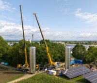 Luftaufnahme zeigt Kräne, die Wärmespeicher bewegen neben einer Freiflächen-PV-Anlage.