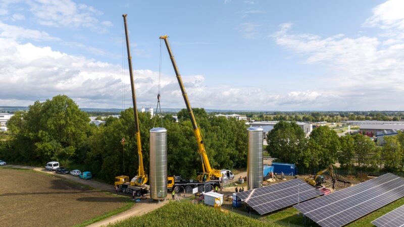 Luftaufnahme zeigt Kräne, die Wärmespeicher bewegen neben einer Freiflächen-PV-Anlage.