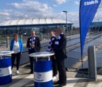 Vier Personen stehen zwischen Bannern des Fußballereins Schalke 04 vor einer Photovoltaikanlage in Gelsenkirchen. Im Hintergrund ist das Fußballstadion.