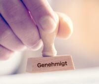 Eine Hand hält einen Holzstempel mit der Aufschrift "genehmigt" - Symbolbild für Genehmigung von Erneuerbare Energien etc.