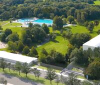Visualisierung zeigt aus der Vogelperspektive weiße Gebäude auf Gelände mit Rasen, Bäumen und Pool - Geothermie-Anlage in München.