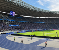 Blick ins Berliner Olympiastadion während eines Fußballspiels: Auf dem Spielfeld stehen Spieler, auf den Tribünen sitzen Zuschauer. Im Vordergrund verläuft die blaue Laufbahn.