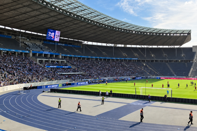 Blick ins Berliner Olympiastadion während eines Fußballspiels: Auf dem Spielfeld stehen Spieler, auf den Tribünen sitzen Zuschauer. Im Vordergrund verläuft die blaue Laufbahn.