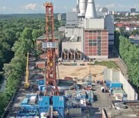 In der Mitte im Vordergrund ein oranger Bohrturm für Tiefengeothermie, links Wald, im Hintergrund ein Kraftwerk