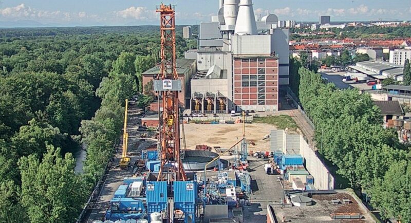 In der Mitte im Vordergrund ein oranger Bohrturm für Tiefengeothermie, links Wald, im Hintergrund ein Kraftwerk